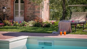 Una piscina con una silla de descanso y una mesa, rodeada por un jardín con flores y un edificio de piedra al fondo.