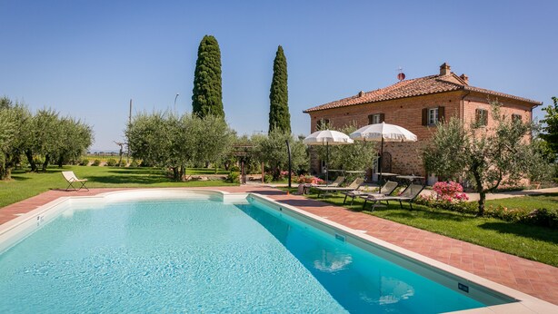 Una piscina rectangular con borde de azulejos, rodeada por un patio de ladrillo. Se observan sillas de descanso y sombrillas al fondo, y una casa de ladrillo con techo de tejas.