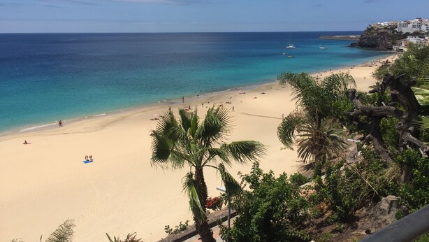 Uma praia com águas azul-claras, areal arenoso e alguns edifícios ao fundo.