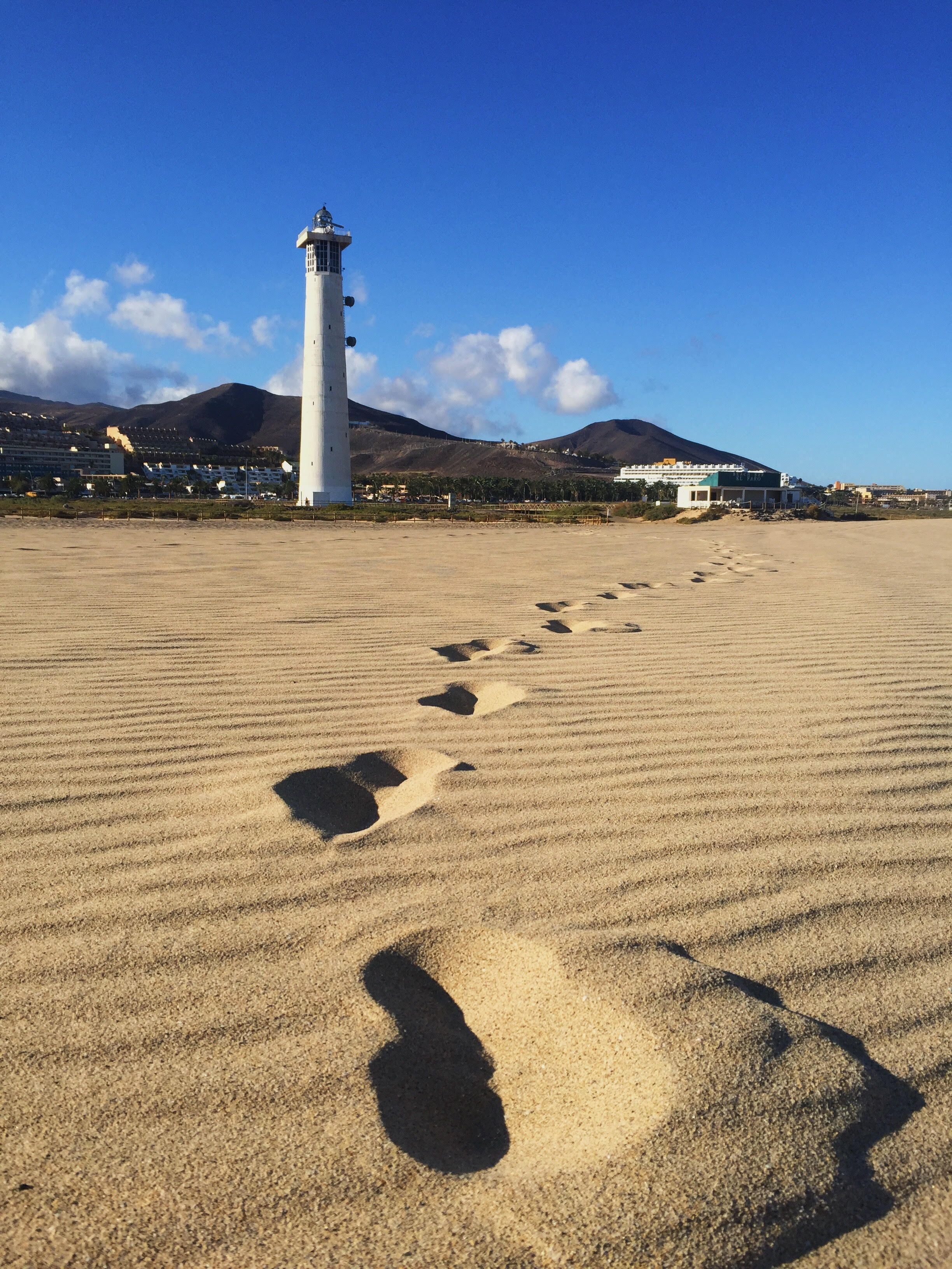 Une plage de sable où l’on peut voir des empreintes de pas menant à un phare blanc.