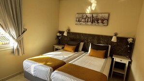 Uma cama de casal com roupa de cama branca e mostarda, duas mesas de cabeceira com abajures, um quadro de uma paisagem urbana e uma janela com cortinas.