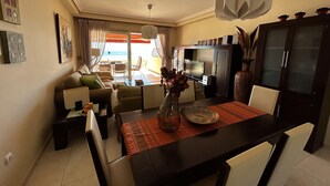Uma sala de estar moderna com um sofá, mesa de centro e mesa de jantar. Há uma televisão, um armário e vista para o exterior.
