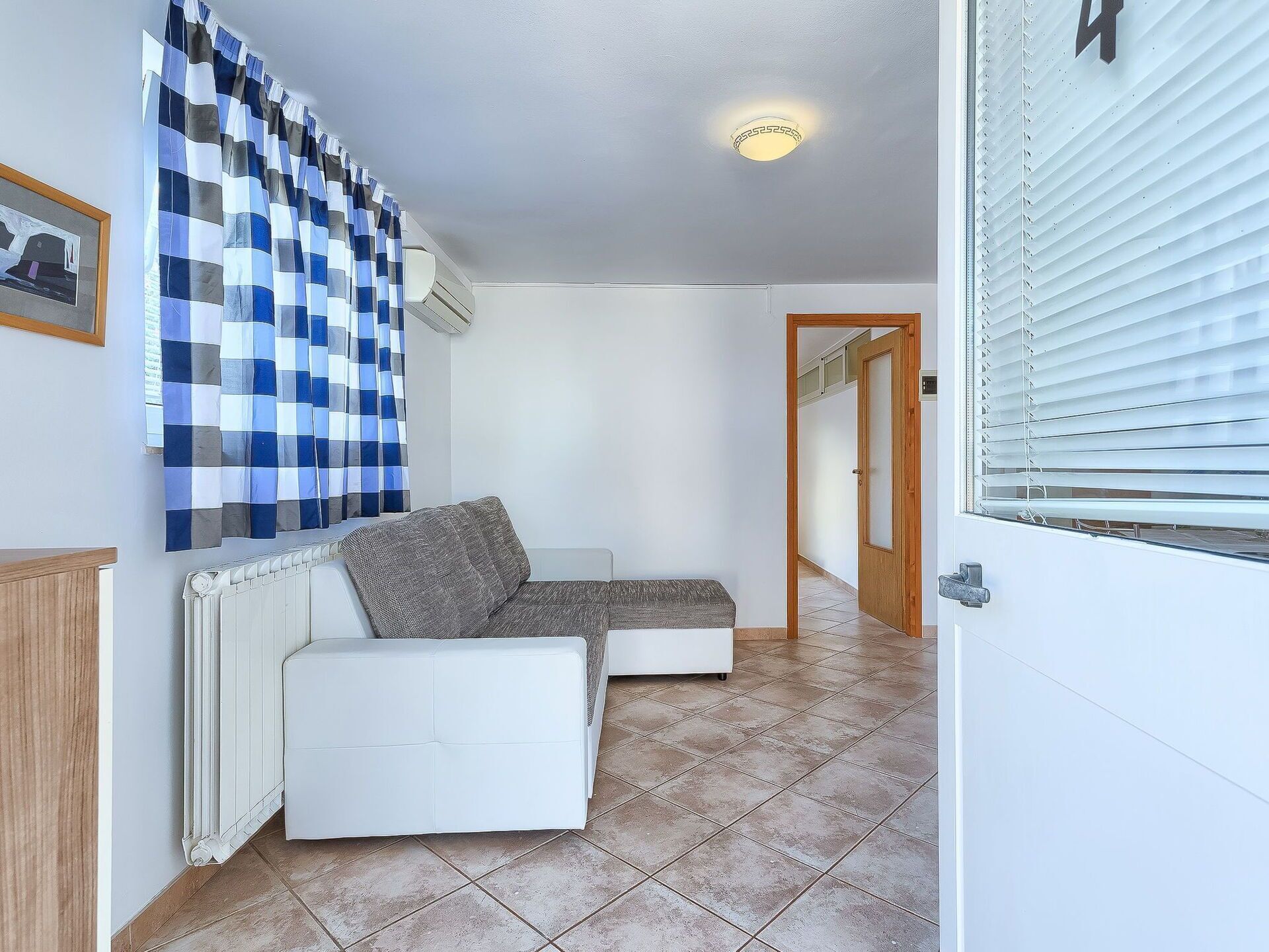 Une pièce avec un canapé blanc, un rideau à carreaux, un radiateur et une porte donnant sur une autre pièce.