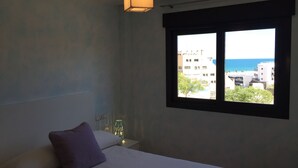 Een slaapkamer met een bed, een raam met uitzicht op gebouwen en de zee, een hanglamp en een nachtkastje met een lamp.