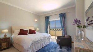 Habitación de hotel con cama, una silla, un escritorio, lámpara, un jarrón con flores y vista al mar por la ventana.