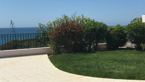 Una terraza pavimentada con vistas al mar, rodeada de vegetación y una barandilla de metal.