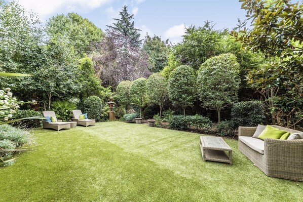 Relax in a serene garden setting surrounded by lush greenery and mature trees, featuring a wicker sofa and lounge chairs.