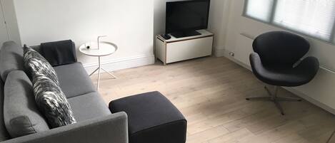 A modern living room with a grey sofa, black ottoman, a small white side table, a black office chair, a TV stand, and a framed picture on the wall.