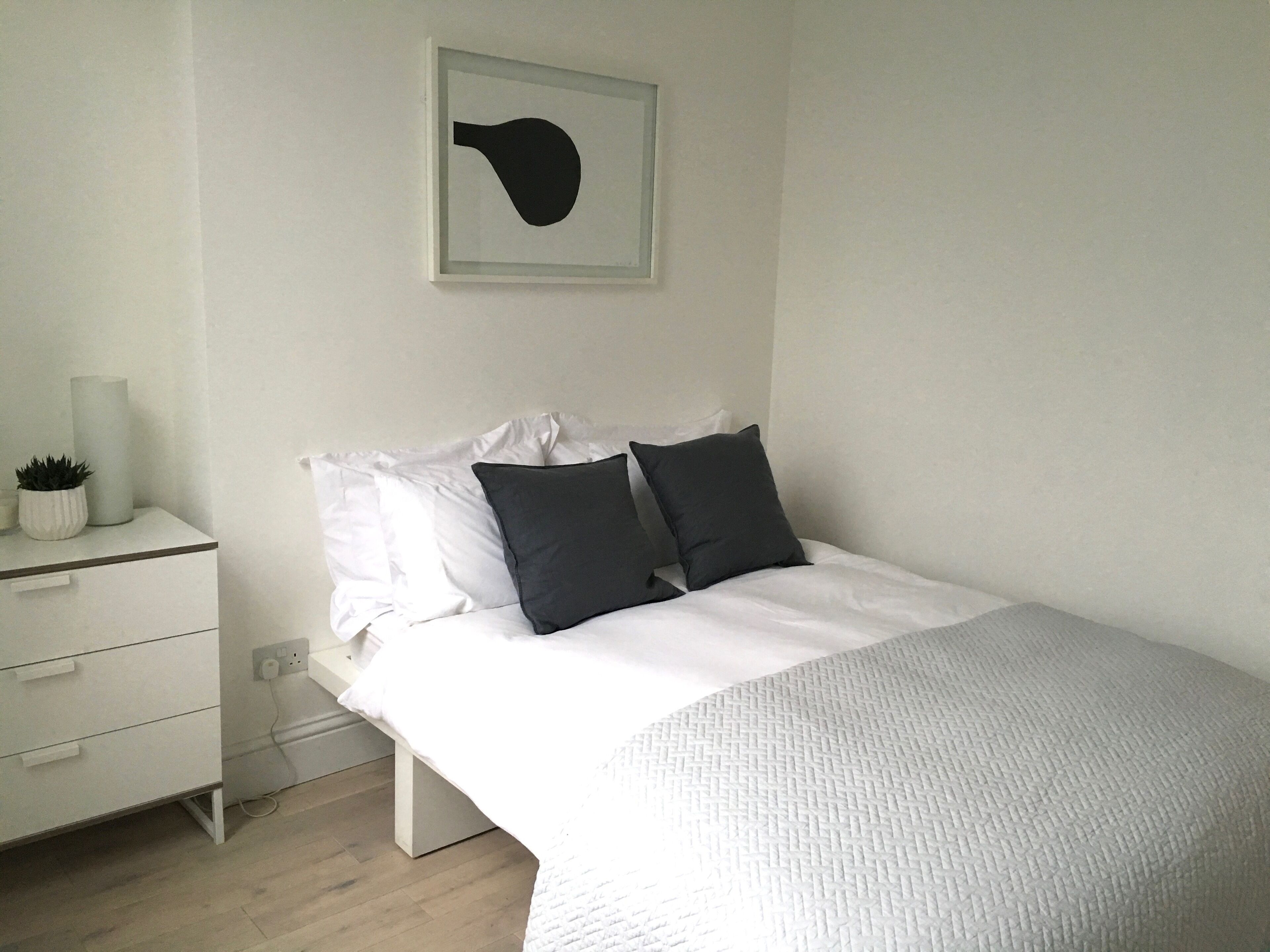 Ein Schlafzimmer mit einem Bett, weißen Nachttischen, einer kleinen Topfpflanze und einem Wandbild.
