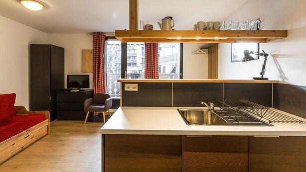 A compact kitchen with a central island, a dining area with a red sofa, and a view of the city through large windows. - Marcel -  an apartment that sleeps 4 guests  in 1 bedroom (Chamonix)