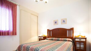 A bedroom with a wooden bed, two bedside tables with lamps, a window with curtains, and two framed pictures on the wall. - Cosy apartment for 2 people with A/C, WIFI, pool, TV and terrace (Calpe)