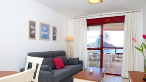 A living room with a grey sofa, a wooden coffee table, and a white chair. There are framed pictures on the wall, a lamp, and a view of the sea through a glass door. - Cosy apartment for 2 people with A/C, WIFI, pool, TV and terrace (Calpe)
