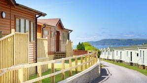 A row of wooden cabins with a stone wall and wooden fence in the foreground, and a clear view of the sea in the background. - Luxury Lodge, South Devon Coast nr Plymouth, 200m to Beach + Indoor Heated Pool, (Nr Plymouth)