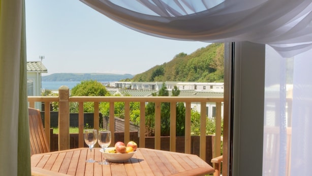 A wooden deck with a table set for two, a bowl of fruit, and two wine glasses. A scenic view of a coastal area with hills and a body of water is visible through the railing. - Luxury Lodge, South Devon Coast nr Plymouth, 200m to Beach + Indoor Heated Pool, (Nr Plymouth)