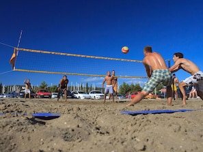 A beach volleyball game with players in shorts and no shirts, a blue net, and cars parked in the background.