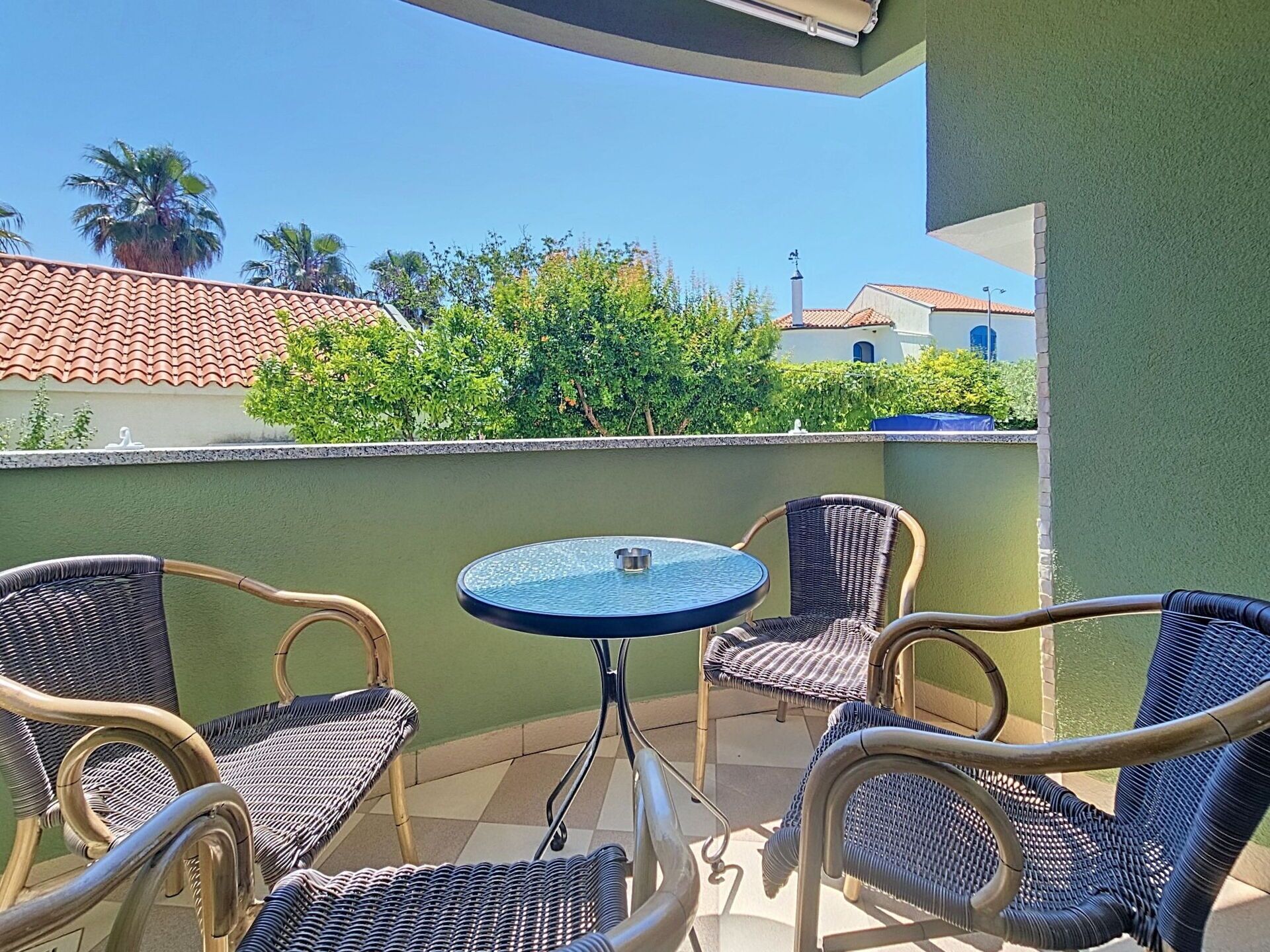 A balcony with wicker chairs and a glass table, overlooking a green wall and a building with a tiled roof.
