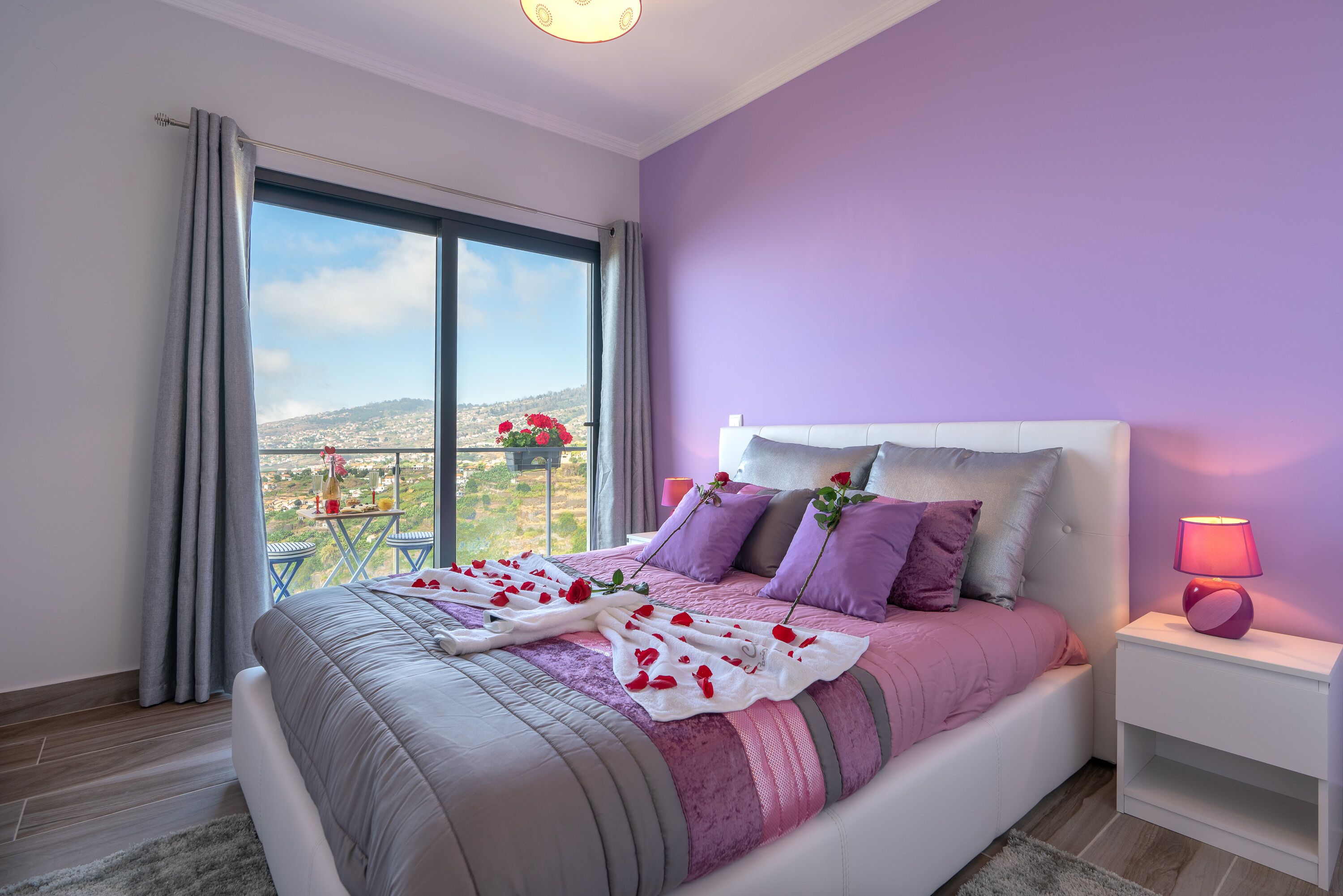 A bedroom with a large bed, a nightstand, a window with curtains, and a view of a cityscape.