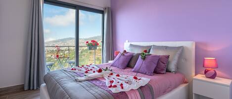 A bedroom with a large bed, a nightstand, a window with curtains, and a view of a cityscape.