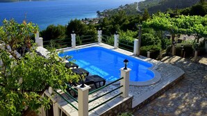A swimming pool with a stone border, surrounded by greenery and overlooking a body of water and hills.