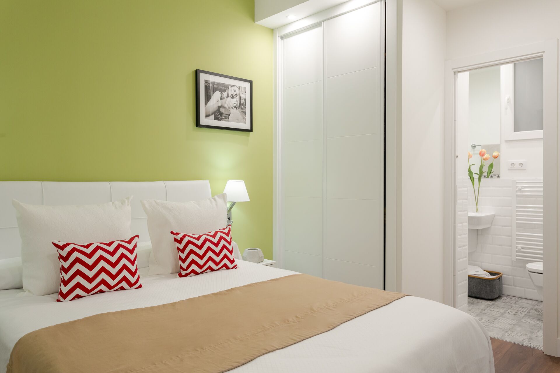 Une chambre d’hôtel avec un lit, une table de chevet, une lampe, un tableau au mur et une salle de bain visible à travers une porte ouverte.