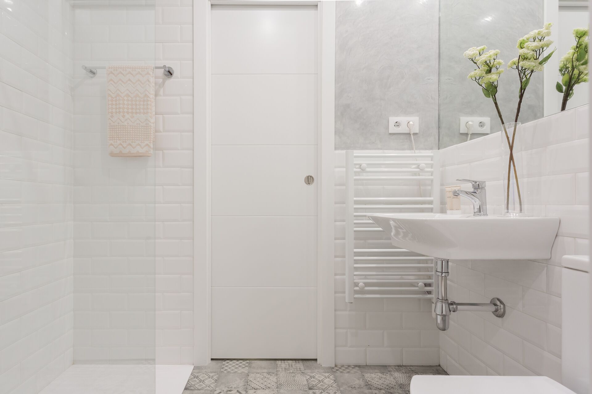 Een moderne badkamer met witte tegels, een douchecabine van glas, een wit wastafelmeubel met spiegel en een handdoekhouder.
