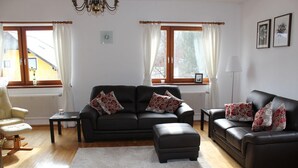 Ein Wohnzimmer mit einer schwarzen Couch, einer kleineren Couch, einem Couchtisch und einem Teppich. Zwei Fenster mit Vorhängen, ein Heizkörper und eine Uhr an der Wand sind zu sehen.