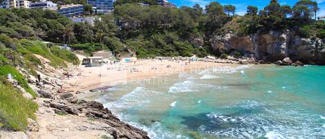 A coastal beach with rocky terrain, clear turquoise waters, and a backdrop of buildings and greenery.