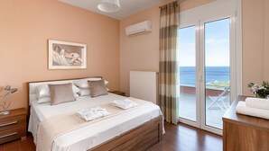 Un dormitorio con cama, mesitas de noche, un cuadro, unidad de aire acondicionado, vista a través de una puerta corrediza y zona de baño con lavamanos y perchero para toallas.