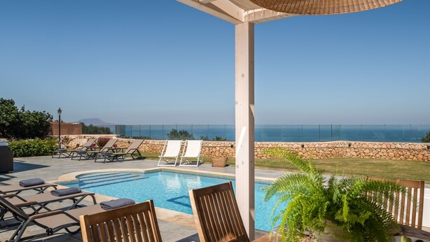 Un área junto a la piscina con sillones de descanso, una mesa con una fuente de fruta, una planta en maceta y vistas al mar.