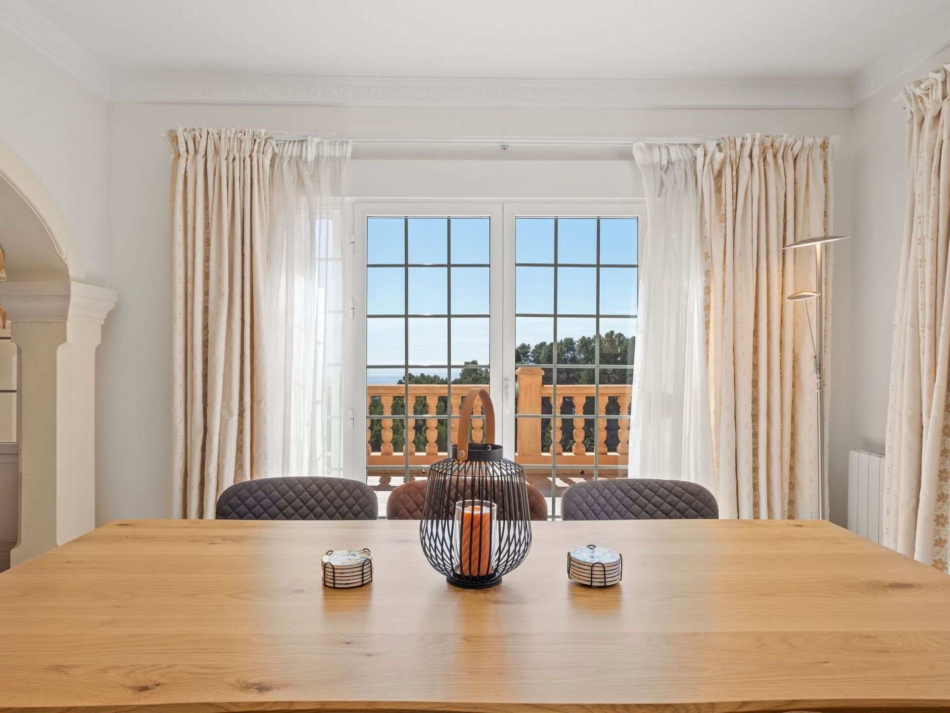 Een eethoek met een houten tafel, twee kandelaars en uitzicht op een balkon met een hekwerk en bomen.