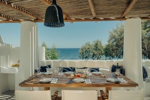 Mesa lista para cuatro personas con vista al mar, pilares blancos y una lámpara colgante.