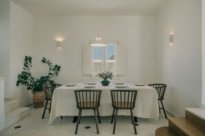 Comedor con mesa blanca dispuesta para cuatro personas, rodeada de sillas, una ventana arriba y una planta a la izquierda.