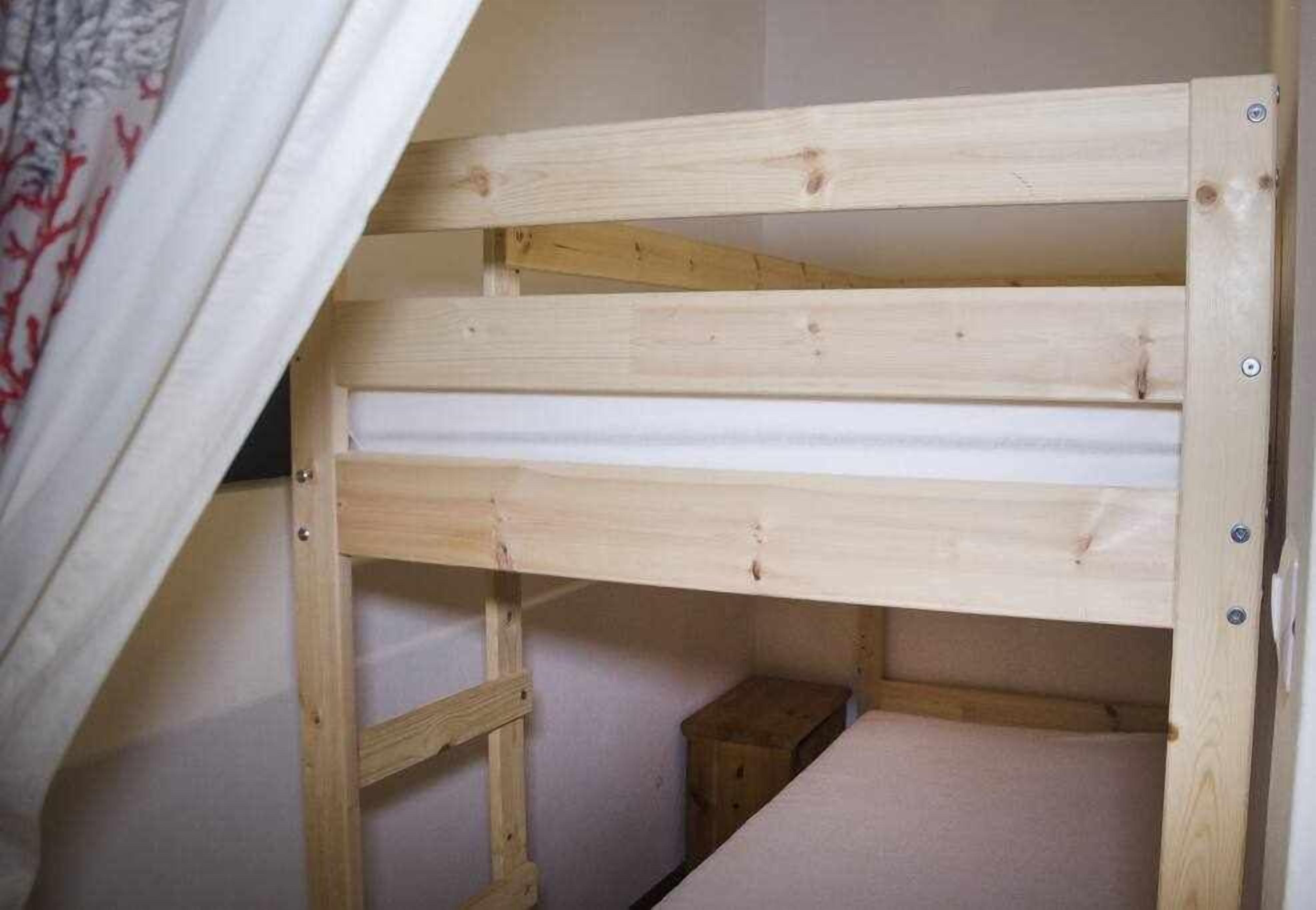 Una cama litera de madera con una escalera, un pequeño cofre de madera y una cortina blanca.