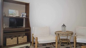 Ein Wohnzimmer mit Fernseher auf einer Holzablage, zwei weiße Stühle, einem Korbtisch mit Laterne und Körben am Boden.
