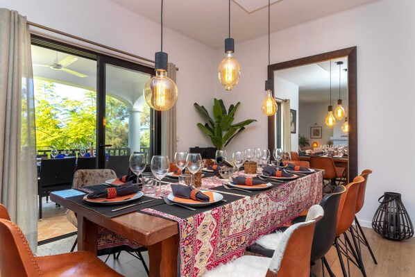 A dining area with a long table set for multiple guests, overhead pendant lighting, and a view of the outdoors.