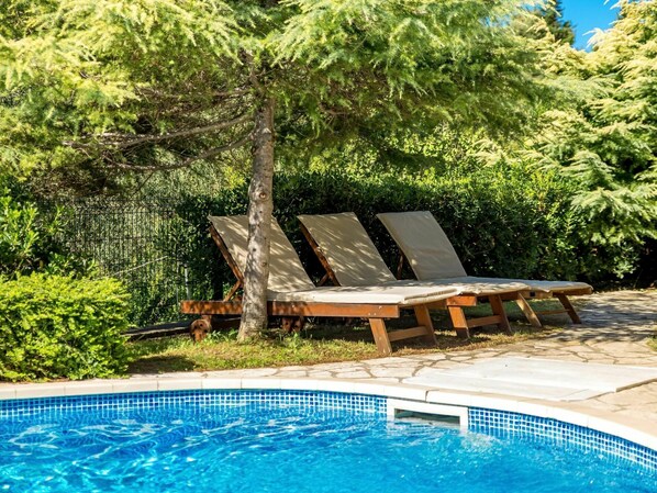 Relax by the serene pool surrounded by lush greenery and shaded by a large tree.