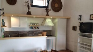 A compact kitchen with a central island, a dining table with a checkered tablecloth, and a television on a shelf. - The Casetta di Merau Sant'Antioco (Sant'Antioco)