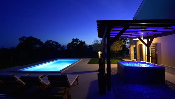 Ein rechteckiger Pool mit Whirlpool unter einer Pergola, umgeben von Liegestühlen und einer kleinen Gartenanlage.