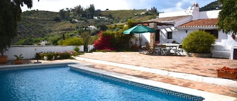Una piscina con borde de azulejos, rodeada por una terraza amueblada con mobiliario exterior y un parasol verde.