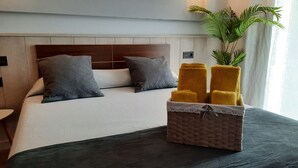 Una habitación de hotel con cama, almohadas grises, una colcha oscura y una cesta de toallas.