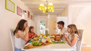Una familia cenando en una mesa blanca, con una araña y rodeada de obras de arte y una escalera.