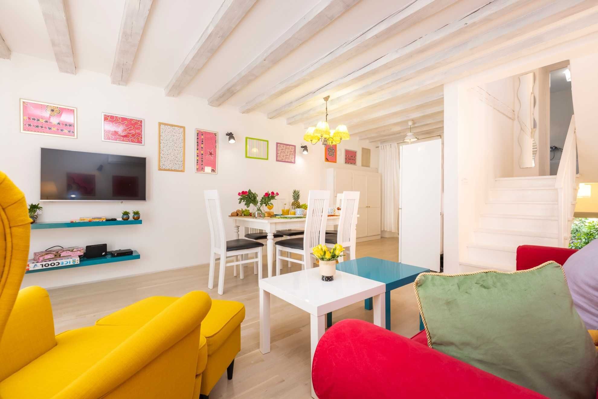 Un salon avec un téléviseur, des canapés jaunes et rouges, un coin repas avec une table et des chaises, et un escalier.