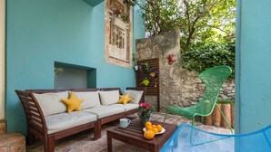 Un rincón exterior acogedor con un sofá de madera, una silla de metal azul y una mesa baja con naranjas y un jarrón de flores.