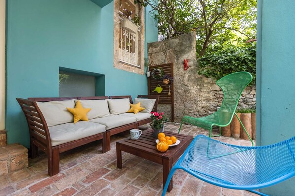 A cozy outdoor seating area with a wooden sofa, a blue metal chair, and a coffee table with oranges and a vase of flowers.