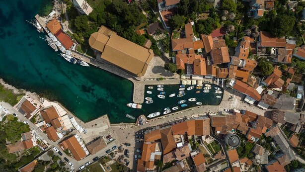 Vista aérea de un pueblo costero con un puerto deportivo, edificios con tejados rojos y aguas azules cristalinas.