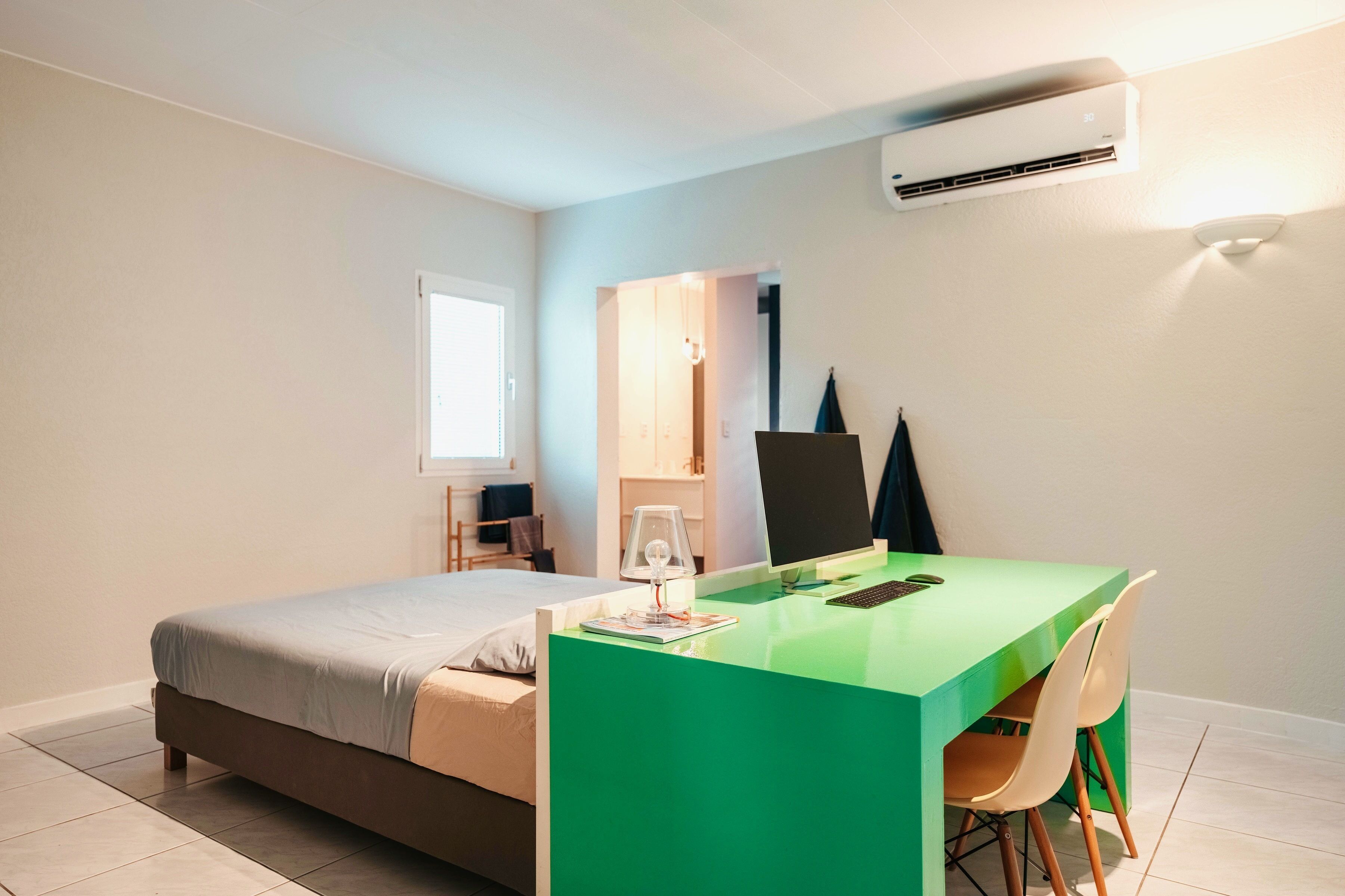 A modern bedroom with a bed, a desk with a chair, a lamp, and an air conditioning unit.