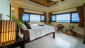 A bedroom with a large bed, a ceiling fan, a view of the ocean, a chair, and a small table with a plant. - White Azure (Ko Pha-ngan)