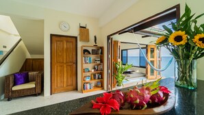 A room with a wooden bookshelf, a wicker sofa, a dining table with flowers, and a view of the outdoors. - White Azure (Ko Pha-ngan)