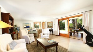 A spacious living room with a sofa, armchairs, a coffee table, and a desk. There is a large window with curtains, a fan, and a view of the outdoors. - Entertainment-packed, Spacious, Peaceful & Convenient (Pollença)