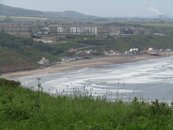 Un pueblo costero con playa, edificios residenciales y una gran instalación industrial al fondo.